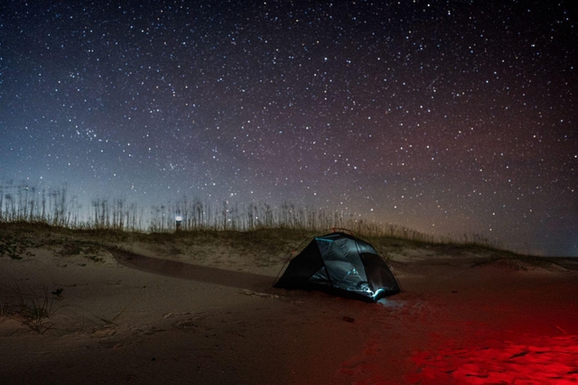 A red light illuminates a tent on the sand, with a star filled sky behind it.
