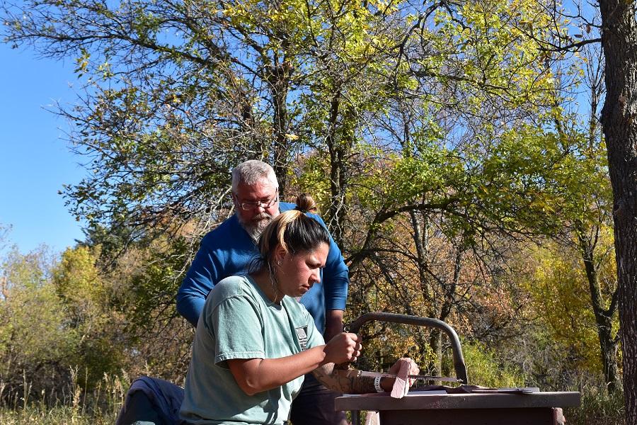 A man watches over a woman as she saws a piece of stone