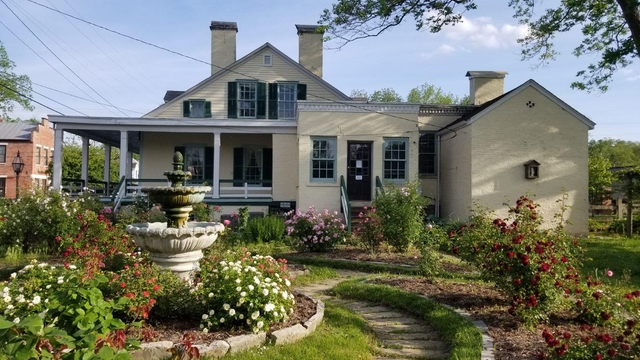 Flower beds with blooming flowers and a fountain, with a cream-colored house behind.