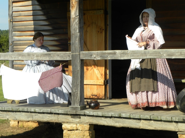 Women in reproduction historic dress sit and stand on porch of old cabin