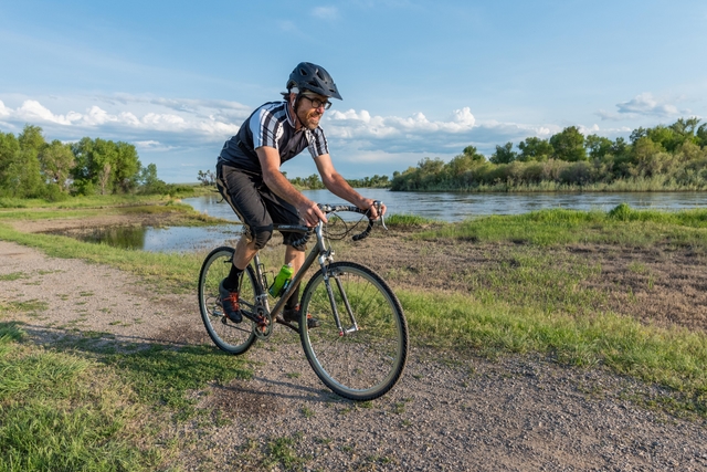 Cyclist biking dirt path along riverfront. Leafy trees and wetlands habitat in distance.