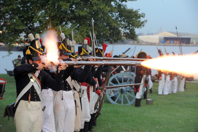 People in 18th century period clothing standing in a line and firing muskets.