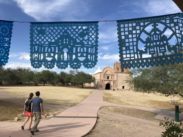 two visitors on trail walking toward church, papel picado in foreground