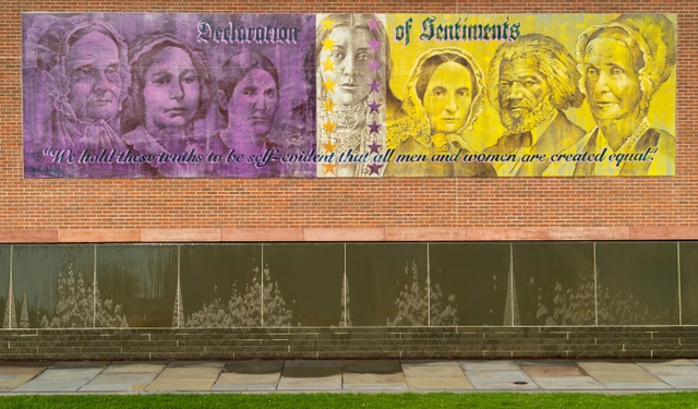 A gold and purple mural featuring faces of historic figures, over a stone wall.