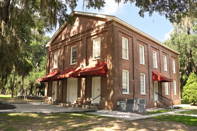 A two story brick building flanked by large live oak trees