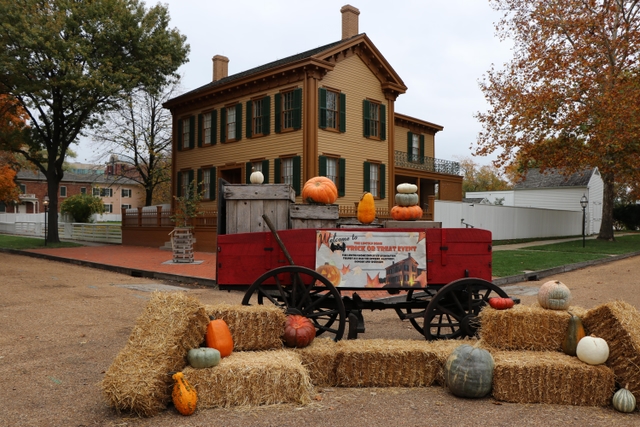 A red cart surrounded by pumpkins and hay bales with the Lincoln Home in the background.