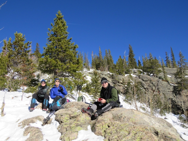Three people are sitting on rocks on a winter trail. There are patches of snow on the ground.