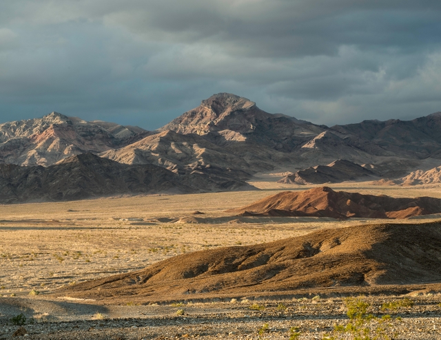 Rugged gray mountains rise over a brown desert landscape under a gray sky.