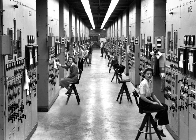 Several women sit on stools along a long hallway full of dials and meters.
