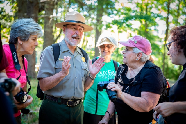 A park ranger in uniform appears to lead a spirited discussion with a group of visitors in a wooded